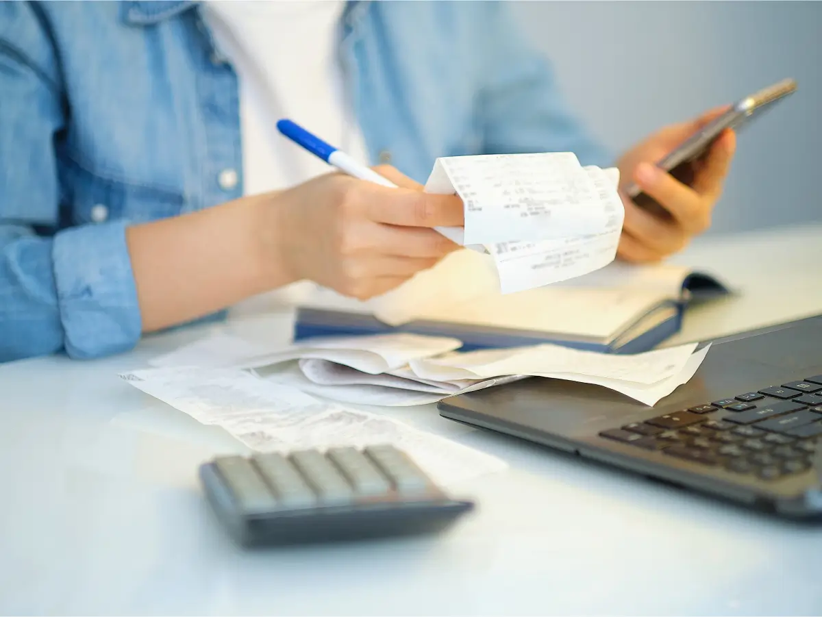 Eine Person sitzt an einem Schreibtisch und überprüft Belege mit einem Stift in der Hand, während sie ein Smartphone benutzt, um eine Haushaltsbuch App zu bedienen. Auf dem Tisch liegen ein Laptop, ein Taschenrechner und weitere Dokumente, was die digitale Verwaltung und Organisation von Finanzen verdeutlicht.